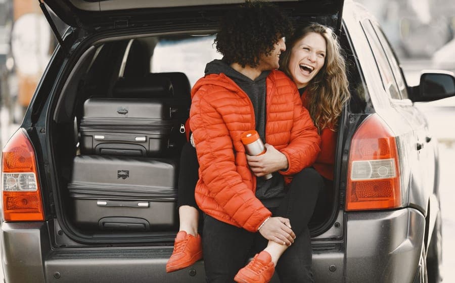 Two people sitting in an open car cargo area with suitcases packed around them